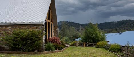 A stone building with a metal roof and wooden beams, surrounded by a grassy area and trees.