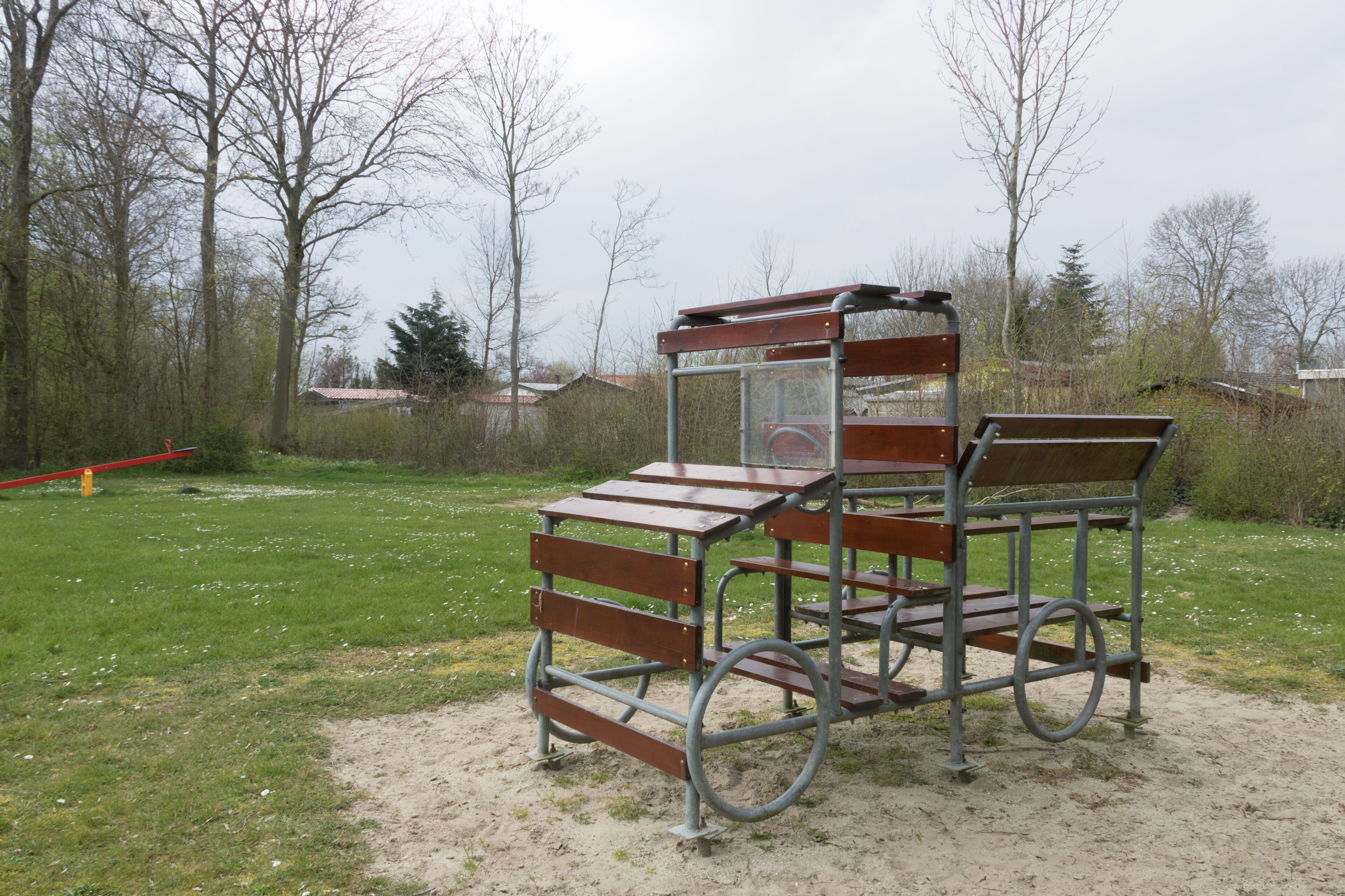 A metal bench with a backrest and a table, situated on a grassy area with trees and a red pole in the background.