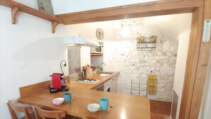 A wooden dining table with chairs, a coffee machine, and a gas stove. A kitchen with a stone wall and a clock is visible in the background.