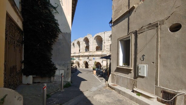 A narrow street with a stone building featuring arched doorways and a small outdoor seating area.