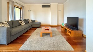 Ein Wohnzimmer mit einem grauen Ecksofa, einem Holztisch, einem Flachbildfernseher auf einem Holzständer und einem Fenster mit Vorhängen.