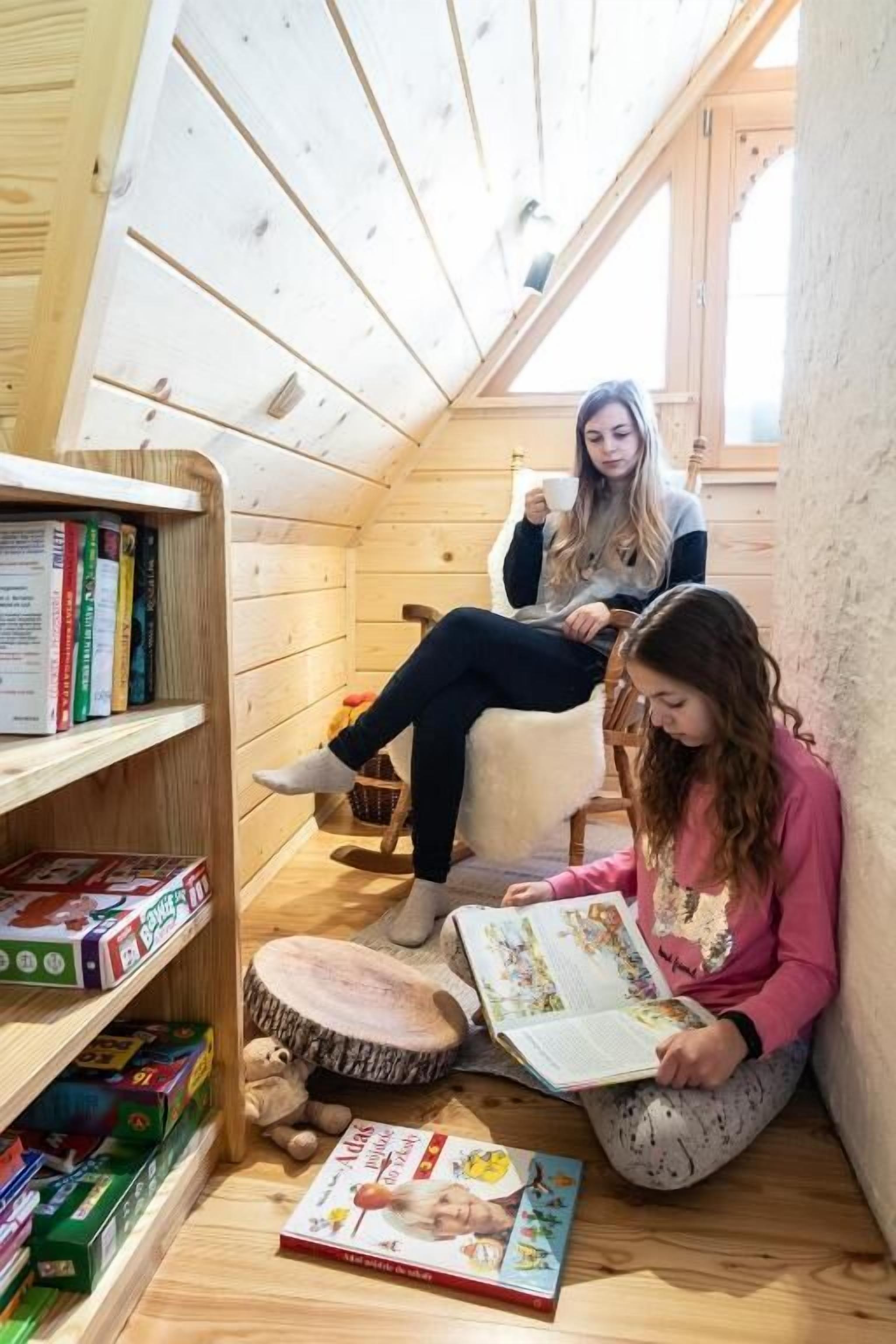 Una habitación acogedora en el ático con paredes de madera y techo inclinado. Dos personas están sentadas en sillas, una leyendo un libro y la otra sosteniendo una taza. Se ve una estantería llena de libros y juegos.