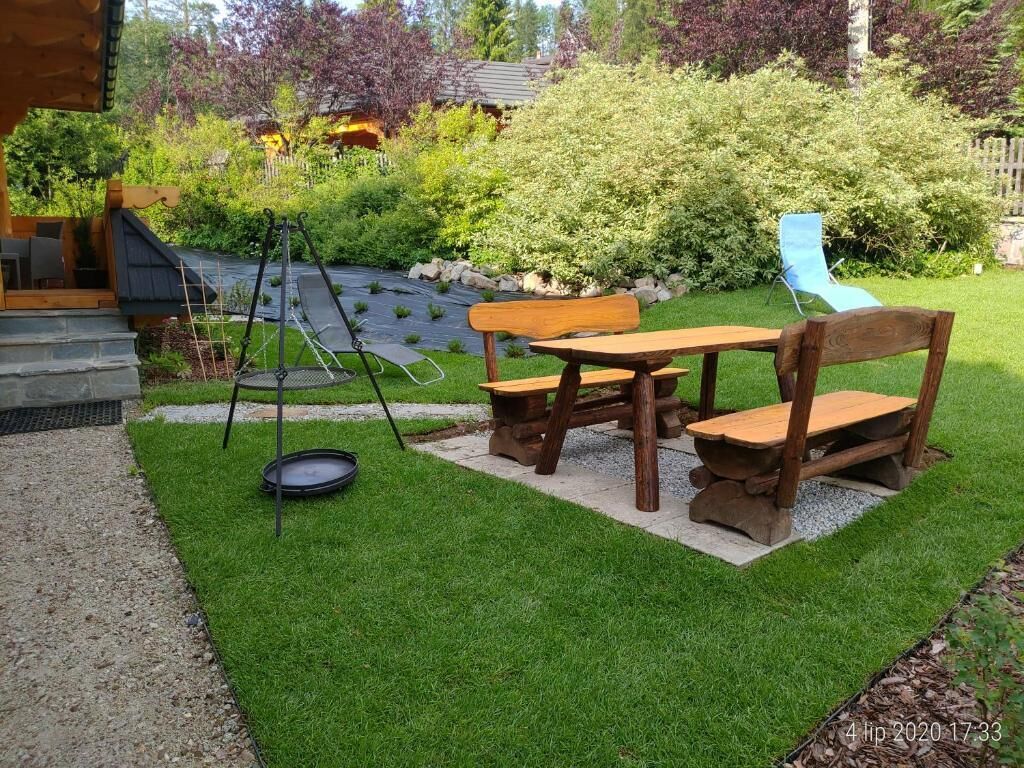 A patio with a wooden table and chairs, a small garden area with a lawn and bushes, and a slide in the background.