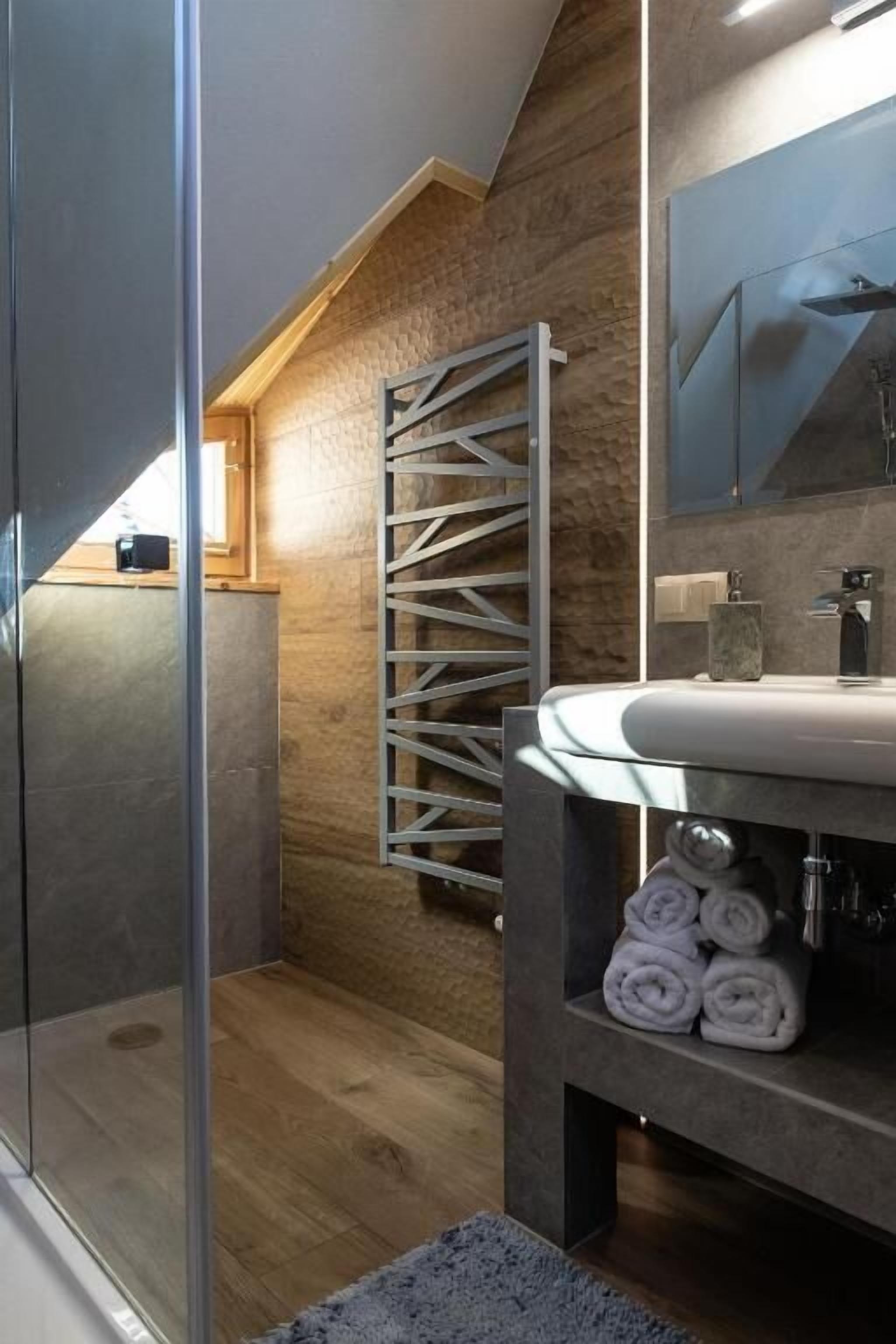 Un baño moderno con techo inclinado de madera, cabina de ducha de vidrio, lavamanos empotrado con encimera de mármol y toallero con toallas dobladas.