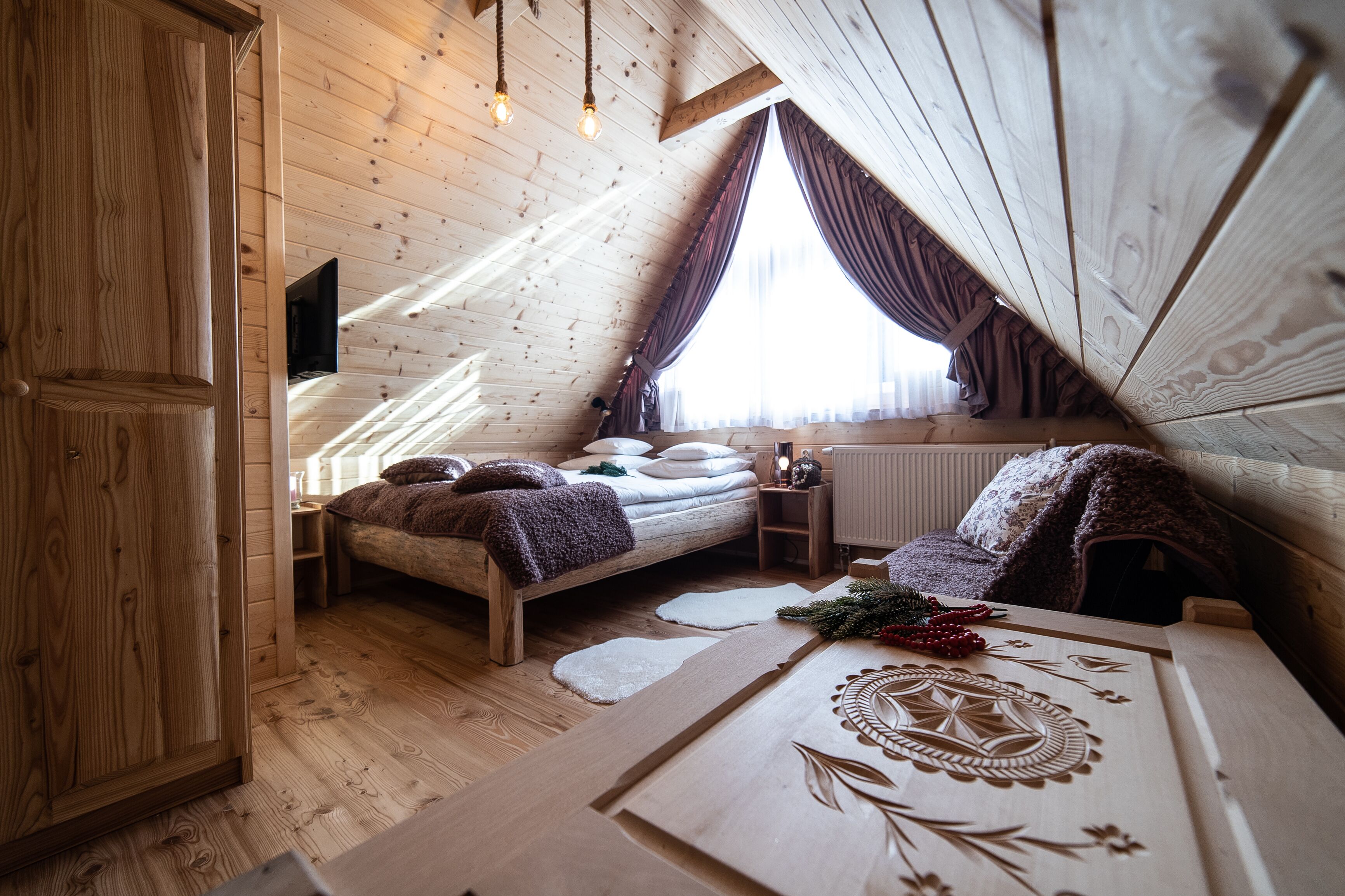 Une chambre confortable située sous les toits, avec un plafond en pente en bois, un lit avec des oreillers et une couverture, une petite table de chevet et une fenêtre avec des rideaux.