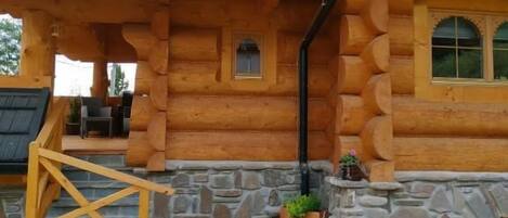 A log cabin with a stone foundation, wooden porch, and a small garden area with potted plants.