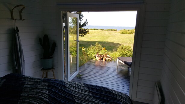 A bedroom with a bed, a cactus, and a door leading to a deck with a view of a grassy field and a body of water. - Panoramic seaview from small island (Omø)