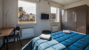 Ein Schlafzimmer mit Bett, Schreibtisch, Stuhl, Fernseher und großen Fenstern mit Blick auf Gebäude.