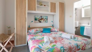 Un dormitorio con cama, una silla de madera, un cuadro y un baño visible a través de una puerta abierta.