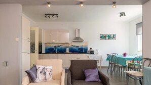 Una sala moderna con un sofá y un sillón, una zona de comedor con una mesa y sillas, y una cocina con armarios blancos y una estufa.