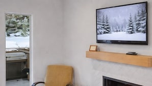 A cozy living room with a fireplace, a comfortable armchair, a television on the wall, and a view of a snowy forest through a window. - Open Floor 71 Vantage Family Condo by RedAwning (Park City)