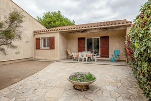 Un patio con suelo de piedra, una jardinera central con plantas, una mesa con sillas y un edificio con techo de tejas y contraventanas de madera.
