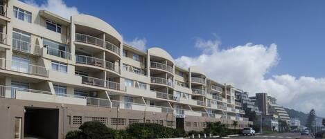 A multi-story residential building with balconies, a parking area, and a clear sky.