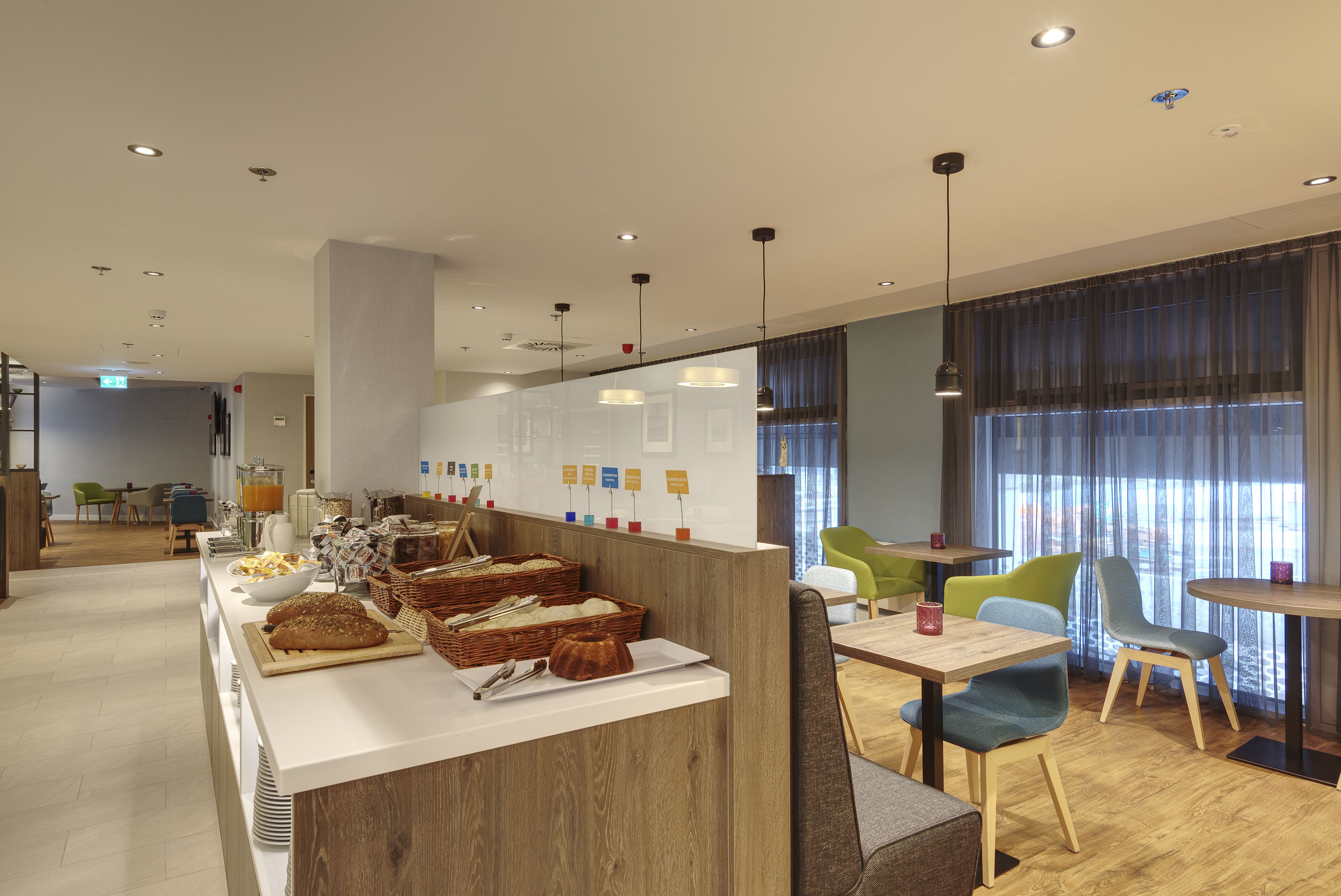 Ein moderner Frühstücksbereich mit Buffet, darunter Brot, Gebäck und eine Obstschale. Es gibt Sitzgelegenheiten mit Stühlen und Tischen, und durch große Fenster ist ein Blick ins Freie möglich.