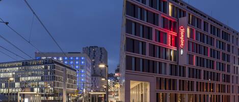 A modern hotel with a prominent red sign, a glass facade, and a tram line in the foreground.