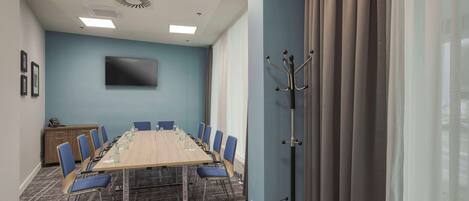 A conference room with a long table, blue chairs, a flat-screen TV, and a coat rack.