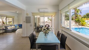 Een moderne eethoek met een glazen tafel en stoelen, uitzicht op een zwembad, en een gezellige zithoek met een bank en kussens.