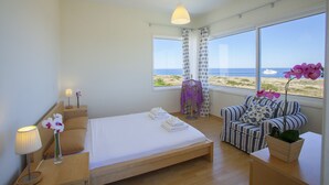 Ett sovrum med en säng, en stol, en lampa och ett fönster med utsikt över stranden och havet.