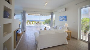Ett rum med en vit soffa, ett litet bord med en lampa, en tv och utsikt över havet genom stora fönster.