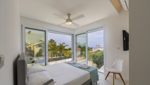 A modern bedroom with a large bed, a ceiling fan, a TV mounted on the wall, and a balcony with a view of the city. - Imagine Your Family Renting This Luxury Contemporary Style Villa in Protaras, Protaras Villa 1487 (Paralimni)