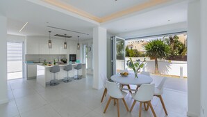A modern kitchen with white cabinets and a central island, a dining area with a round table and four chairs, and a large window offering a view of palm trees and a building. - Imagine Your Family Renting This Luxury Contemporary Style Villa in Protaras, Protaras Villa 1487 (Paralimni)