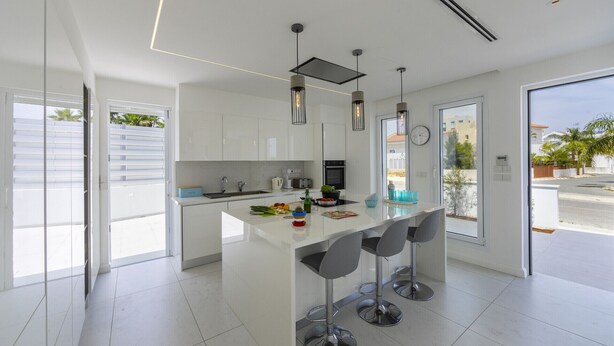 A modern kitchen with white cabinetry, a central island with bar stools, pendant lighting, and a view of the outside. - Imagine Your Family Renting This Luxury Contemporary Style Villa in Protaras, Protaras Villa 1487 (Paralimni)