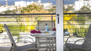 Ein Balkon mit einem für zwei Personen gedecktem Tisch, mit einer Flasche und Gläsern, und Blick auf das Meer und Gebäude im Hintergrund.