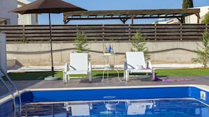 Et poolområde med to hvide liggestole, et bord med flasker og en pergola med en brun parasol.