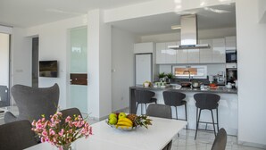 Eine moderne Küche mit weißen Schränken, einem Edelstahlkühlschrank und einer Kaffeemaschine. Es gibt einen Essbereich mit einem weißen Tisch und Stühlen, und auf dem Tisch steht eine Blumenvase.