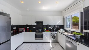 A modern kitchen with white cabinets, black countertops, a built-in oven, a washing machine, and a window with a view of greenery. - Your Dream 5-star Escape: Protaras Villa 1483 (Paralimni)