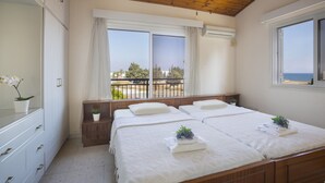 Une chambre avec un plafond en bois, deux fenêtres avec des rideaux, un lit avec une literie blanche, une table de chevet avec une plante, et une vue sur la mer.