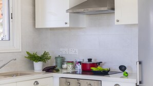 Une cuisine avec des meubles blancs, une cuisinière et un évier. On peut voir des légumes sur le plan de travail et une plante sur la gauche.