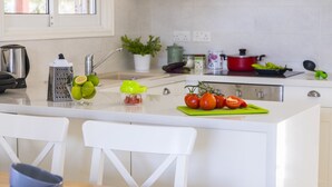 Une cuisine moderne avec un îlot central blanc, une bouilloire, une râpe, un bol de citrons verts et une planche à découper avec des tomates.
