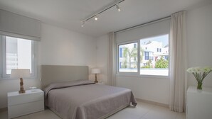 Ein modernes Schlafzimmer mit einem großen Bett, Nachttischlampen, einem Beistelltisch und einem Fenster mit Blick auf Gebäude und Grünflächen.