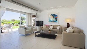 Ein modernes Wohnzimmer mit einer Couch, zwei Sesseln, einem Couchtisch und einem Fernseher. An der Wand hängt ein Gemälde und das Fenster bietet Blick auf Grünflächen.