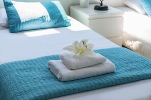 Een net opgemaakt bed met een witte deken, een teal geborduurd sprei en een witte handdoek met een bloem erop.