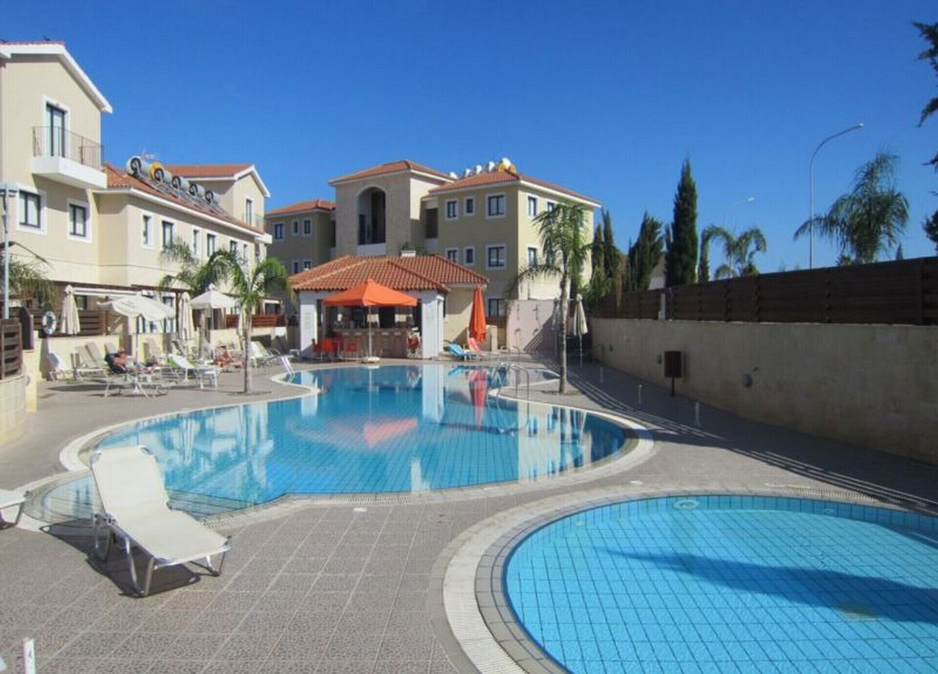 Una piscina con sillones de descanso y sombrillas, rodeada de edificios de estilo mediterráneo.