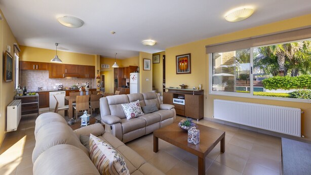 A modern living room with a beige sofa, a coffee table, and a dining area with wooden cabinets and a kitchen in the background. - At Last You can Rent the Perfect Luxury Villa in Larnaca, Larnaca Villa 1392 (Pervolia)