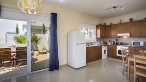 A modern kitchen with a refrigerator, microwave, stove, and washing machine. There is a dining area with a table and chairs, and a large window with curtains. - The Secret to Enjoying Your 5 Star Villa Close to the Beach, Protaras Villa 1484 (Paralimni)