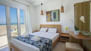 Een hotelkamer met een groot bed, een houten hoofdbord, een nachtkastje met een lamp, een klein bureau met een stoel, en een groot raam met uitzicht op het strand.
