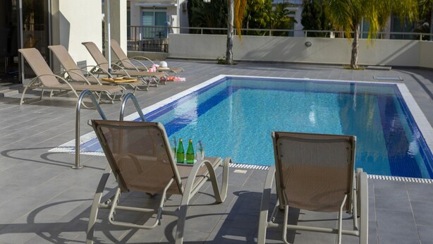 A swimming pool with lounge chairs and umbrellas, surrounded by buildings and palm trees. - Rent Your 5-star Villa With Private Pool Protaras Villa 1525 (Paralimni)