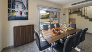 A modern dining area with a wooden table, black chairs, and a fruit bowl. A painting of a rainy street scene is on the wall. - Rent Your 5-star Villa With Private Pool Protaras Villa 1525 (Paralimni)