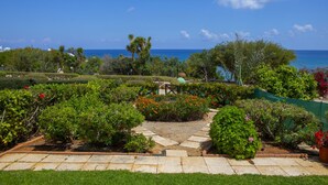 A landscaped garden with a stone pathway leading to a seating area, surrounded by various plants and trees, with a view of the ocean. - Seaside Serenity: Protaras Villa 1515 (Paralimni)