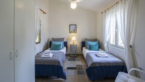 A bedroom with two beds, a ceiling fan, a nightstand with a lamp, and a window with curtains. - Seaside Serenity: Protaras Villa 1515 (Paralimni)