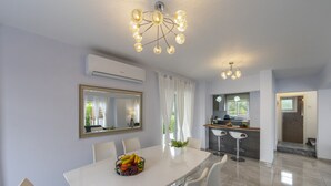A modern dining area with a white table, chairs, and a fruit bowl. There is a kitchen in the background with a bar and a large mirror. - Your Beautiful Holiday Villa Minutes From the Beach, Protaras Villa 1492 (Paralimni)