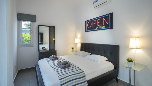 A hotel room with a bed, a mirror, a bedside table with a lamp, and a sign indicating the room is open 24 hours. - Luxury Holiday Apartment, Protaras Apartment 1417 (Paralimni)