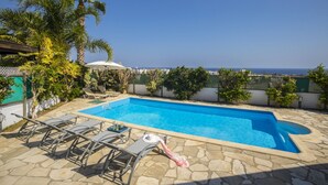 A rectangular pool with a stone border, surrounded by lounge chairs and an umbrella, with a view of the sea and buildings in the background.