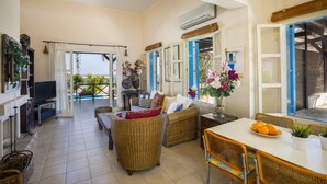 A spacious living area with wicker furniture, a dining table with chairs, and a view of the sea through open doors.