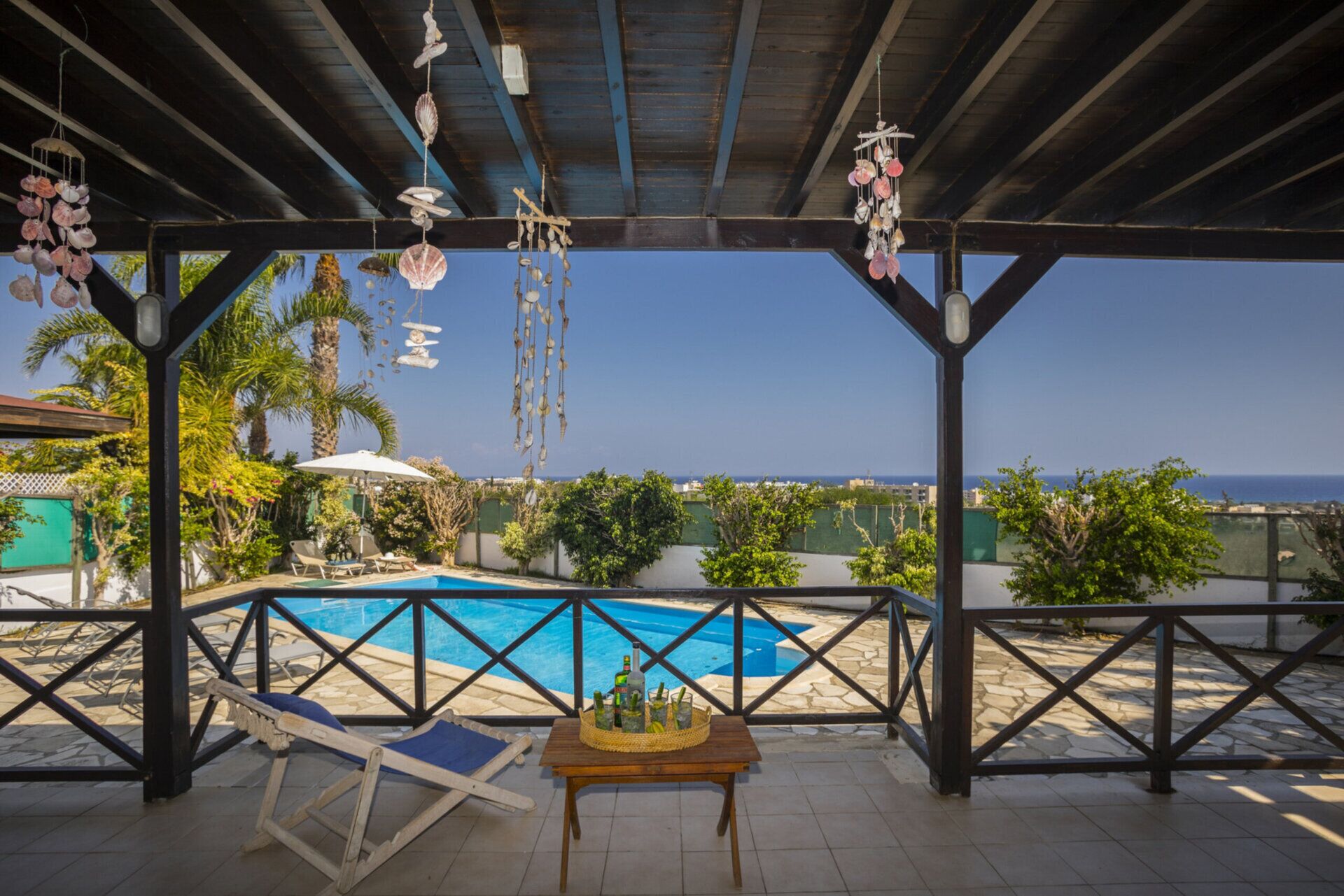 A covered outdoor pool area with a view of the sea, lounge chairs, and a table with bottles.
