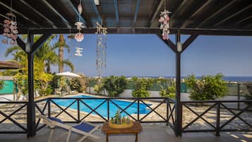 A covered outdoor pool area with a view of the sea, lounge chairs, and a table with bottles.